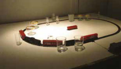 Cadu. Partitura III, 2012. Tren eléctrico, barras de metal, tapa de madera, vasos y botellas de vidrio. Dimensiones variables.